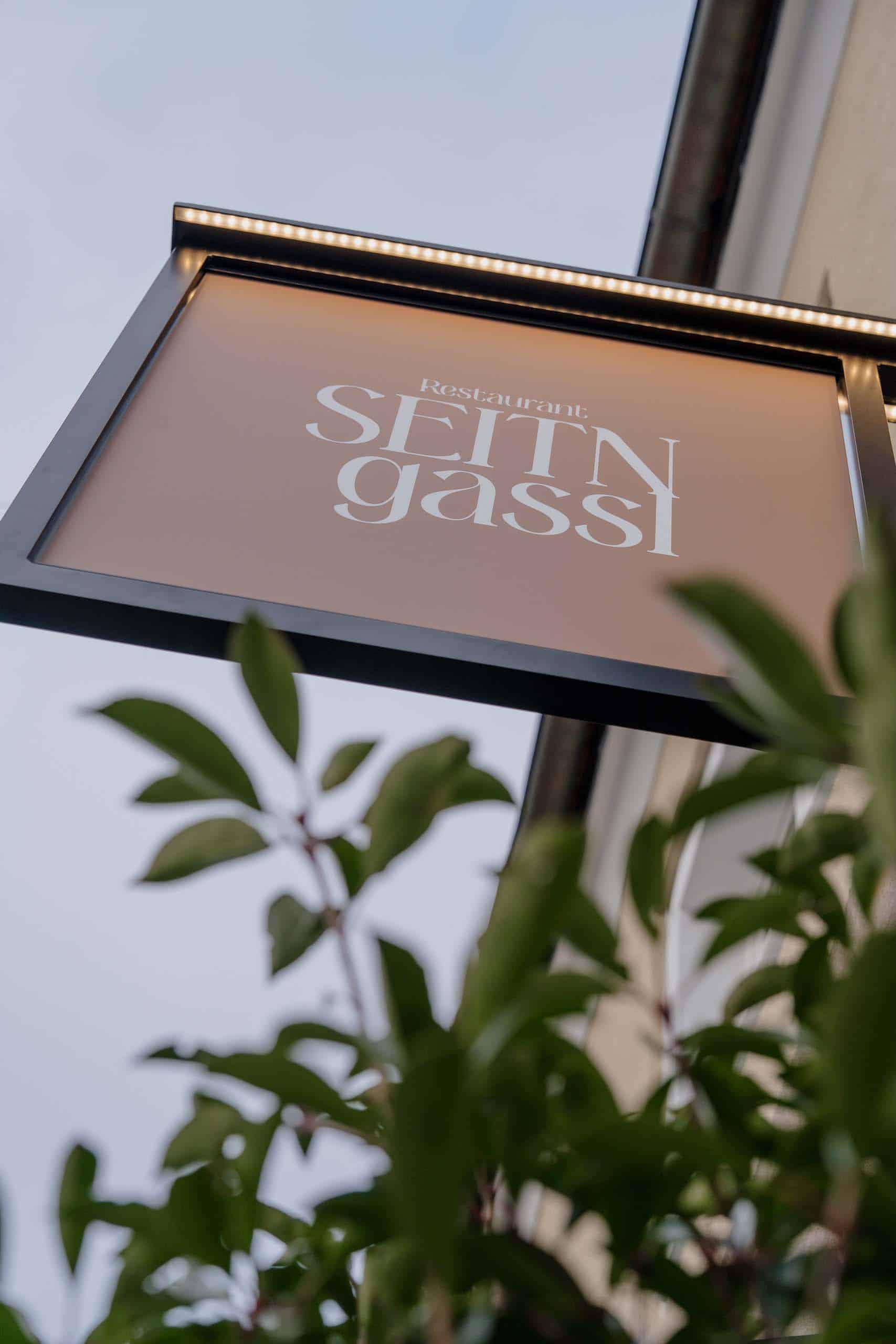 Schild des Restaurants Seit'n Gassl, ideale Hochzeitslocation, umgeben von Pflanzen für romantische Feiern im Freien.