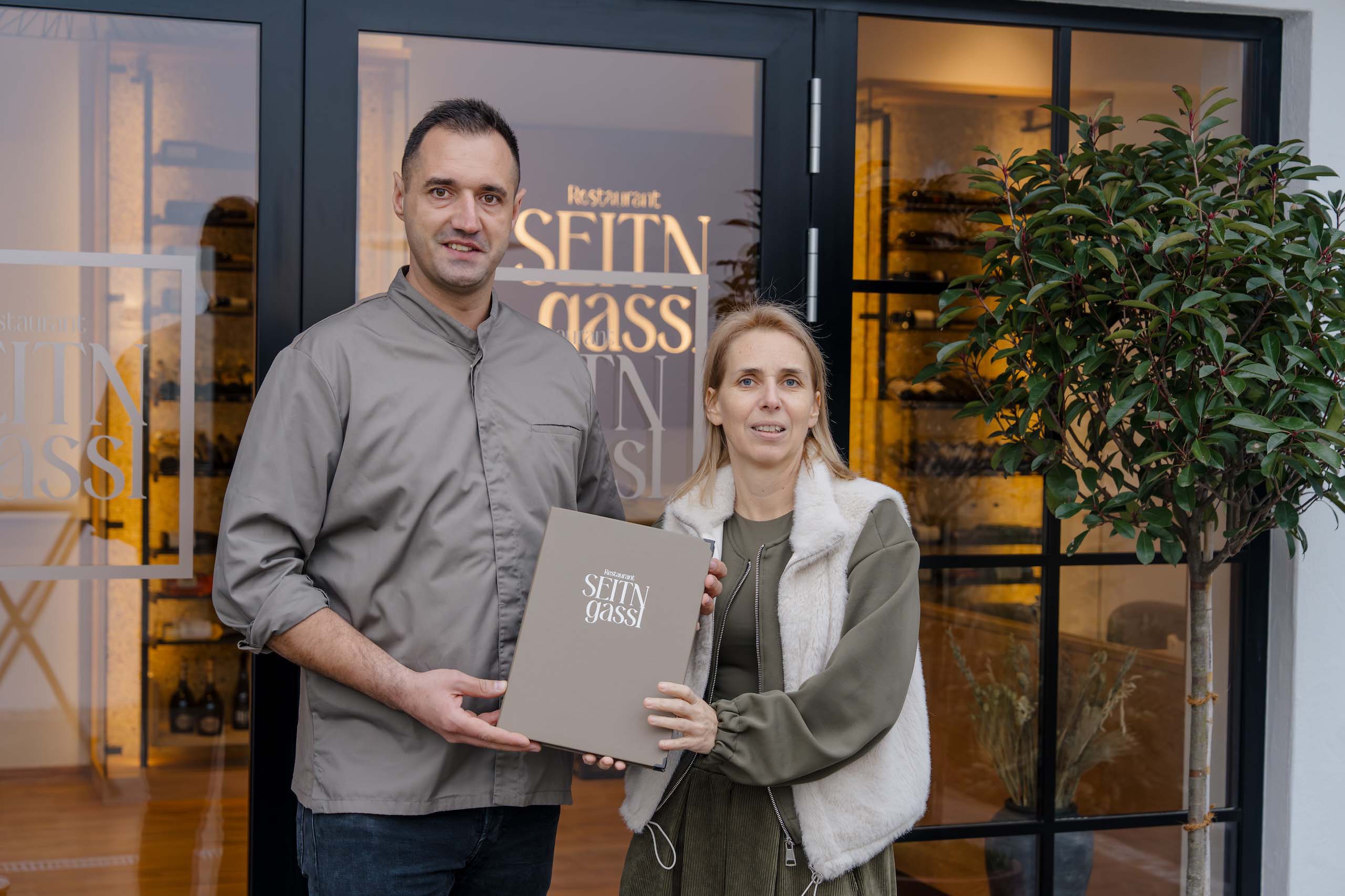 Paar vor stilvoller Hochzeitslocation, hält Schild „Seiten-Gasse“; perfekte Kulisse für Ihren Hochzeitstraum.