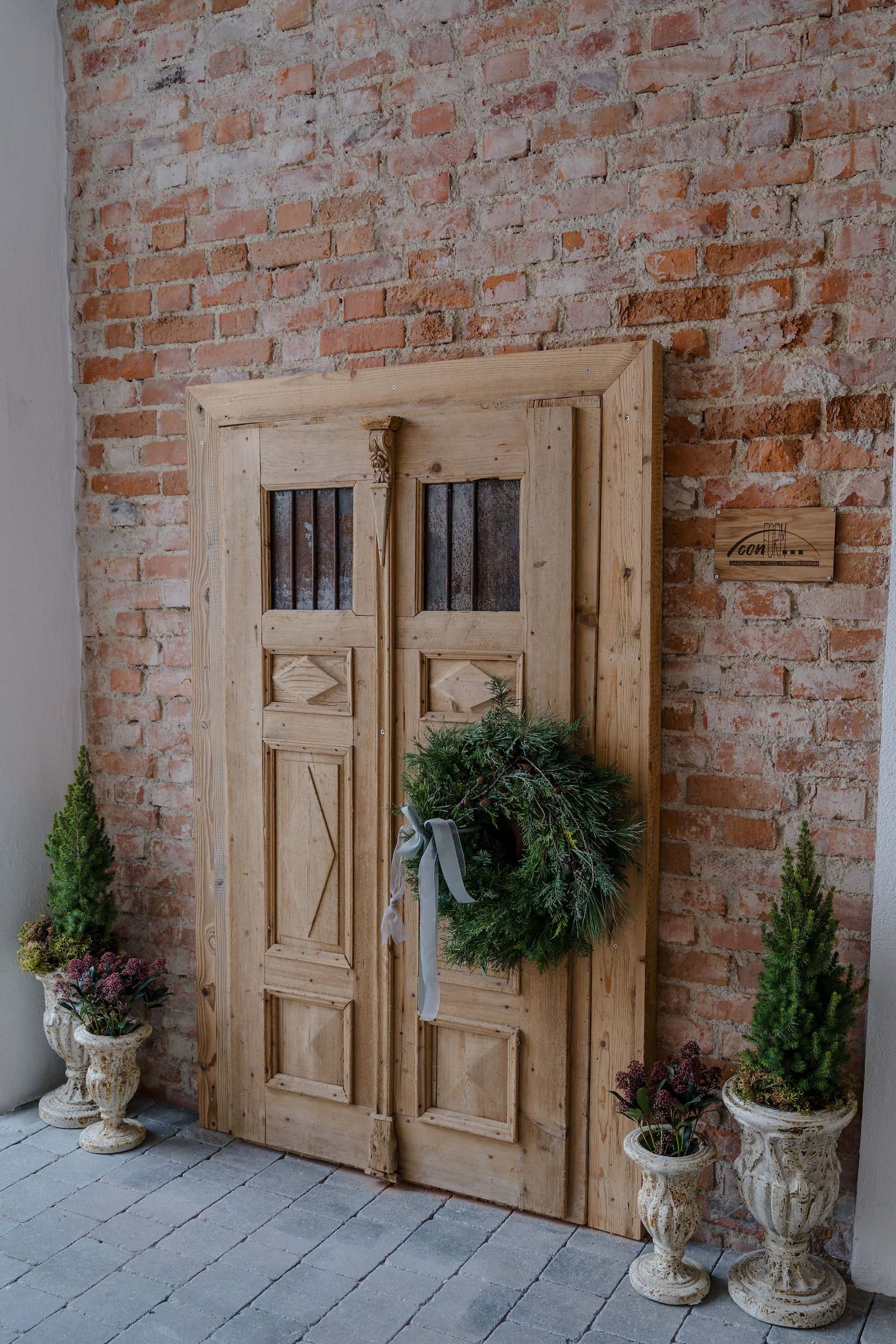 Rustikale Holztür mit Kranz und Pflanzen in antiken Töpfen, ideal für romantische Hochzeitslocation vor Ziegelwand.