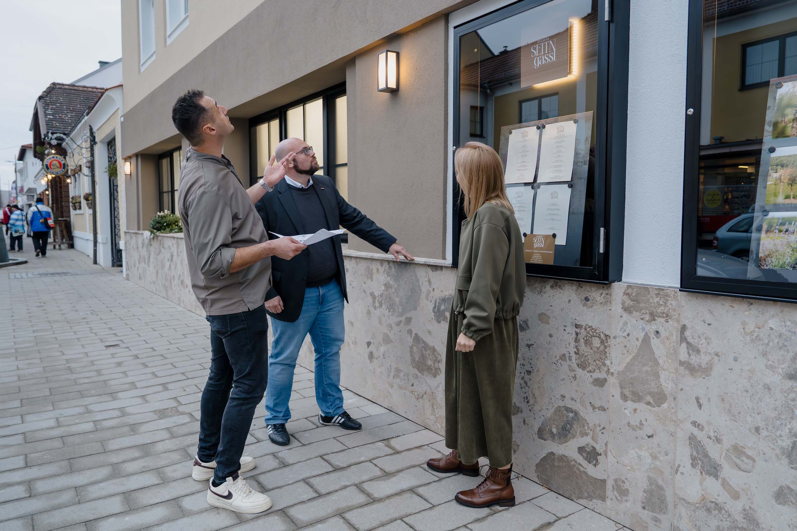 Drei Personen besichtigen potenzielle Hochzeitslocation im Freien, während sie Baupläne und Menüangebote studieren.