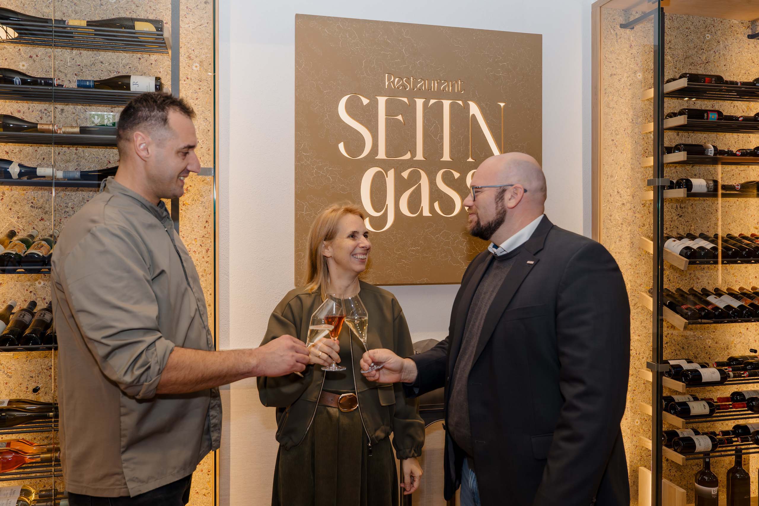 Drei Personen stoßen in einem eleganten Restaurant auf eine Hochzeit an, umgeben von Weinregalen, perfekte Hochzeitslocation.