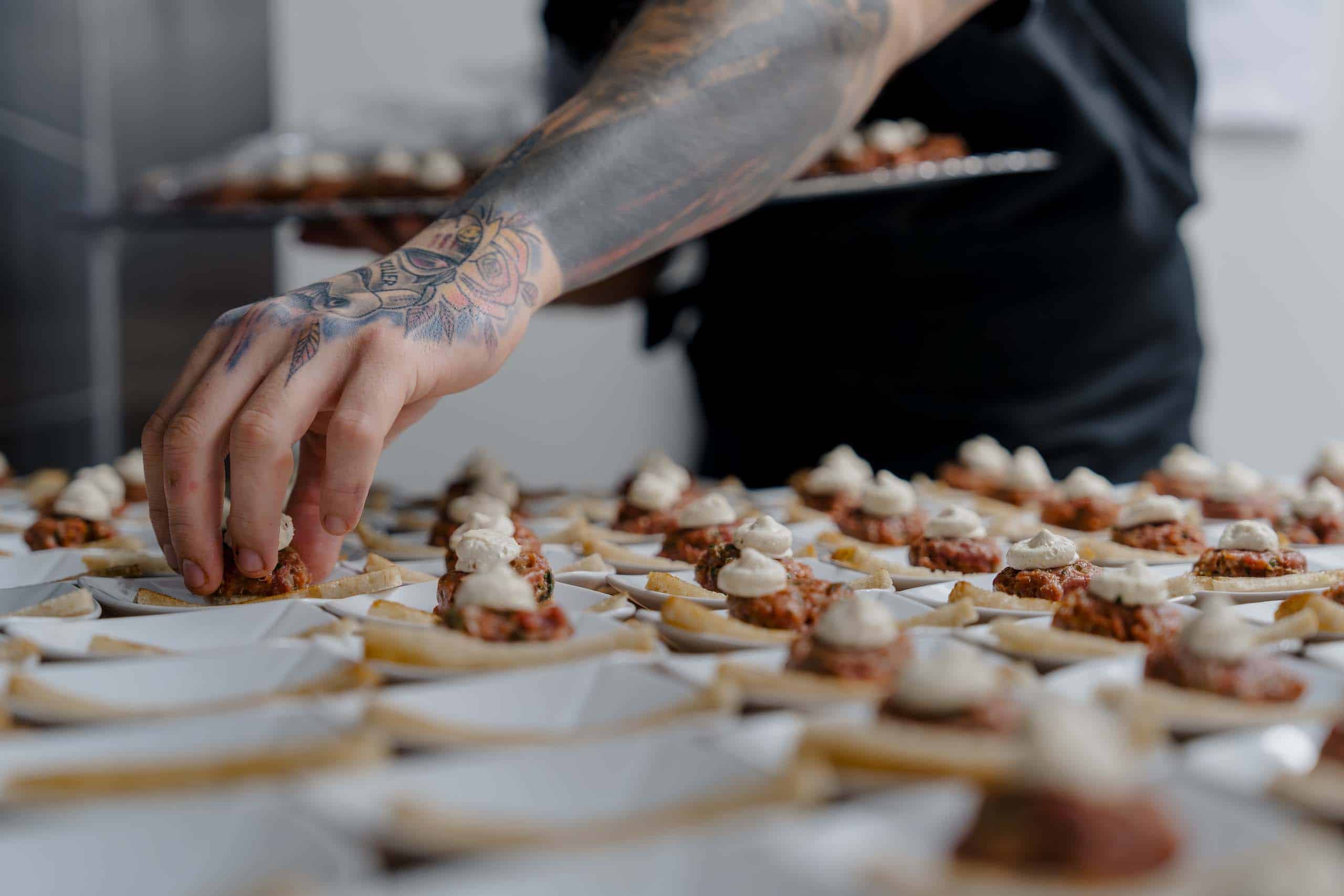 Tattooierte Hand arrangiert kunstvoll Häppchen auf Tablett, perfekte Vorspeisen für eine stilvolle Hochzeitsfeier.