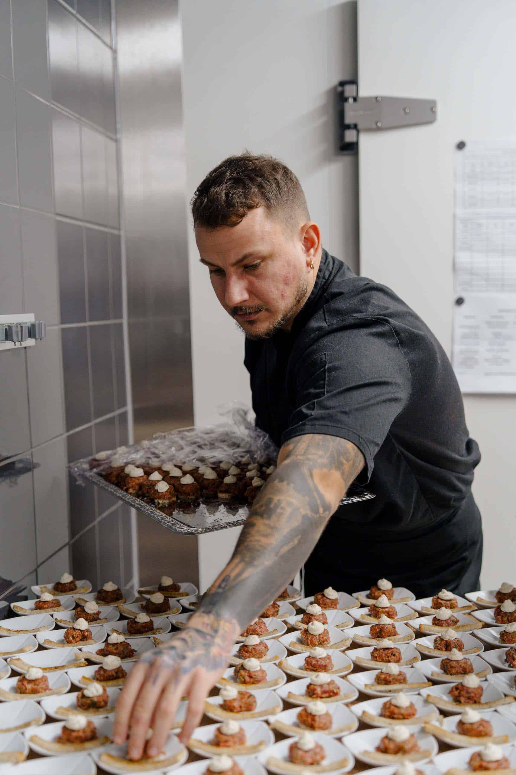 Tatowierter Koch arrangiert kunstvolle Vorspeisen für Hochzeitsbuffet, perfekte Hochzeitslocation für kulinarische Erlebnisse.
