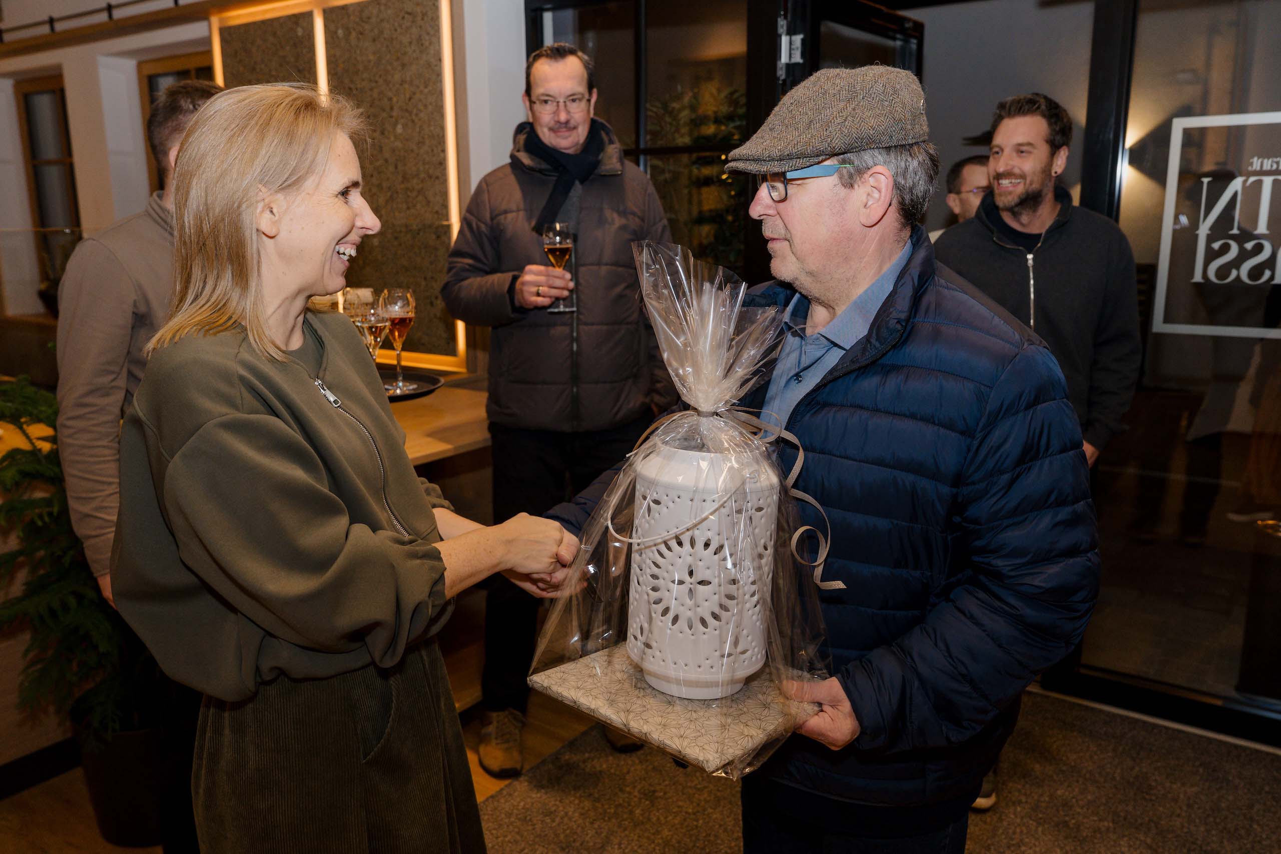 Person überreicht Geschenk bei festlicher Hochzeitslocation, umgeben von Gästen mit Getränken, warme und freundliche Atmosphäre.