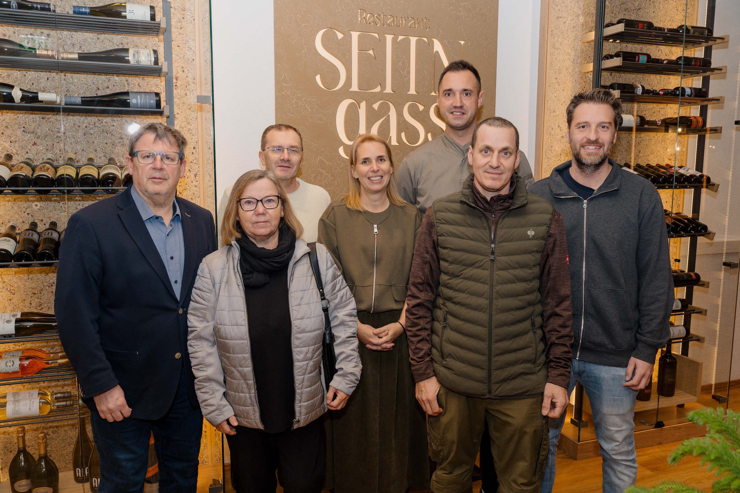Gruppe von Menschen posiert lächelnd in einem stilvollen Restaurant mit Weinauswahl, ideal für Hochzeitsfeiern.