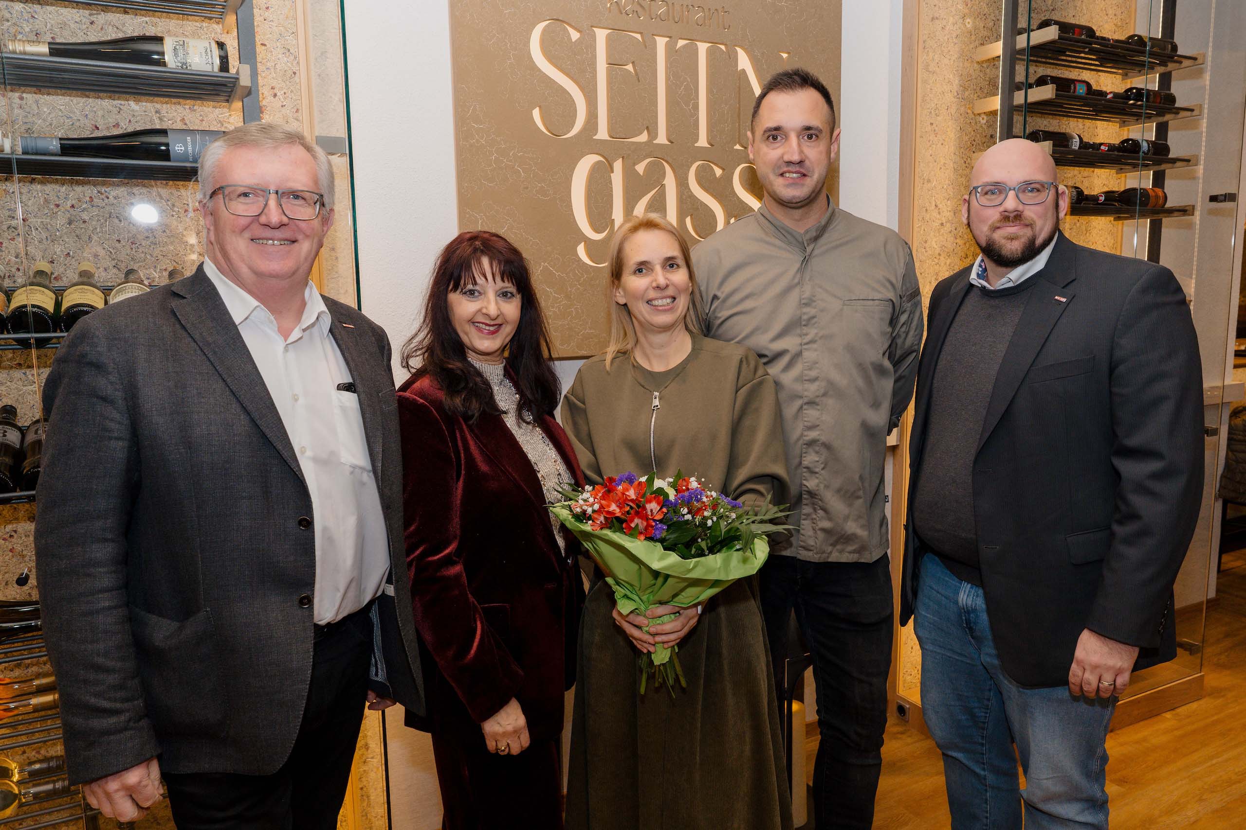 Gruppe von fünf lächelnden Menschen vor dem Schild eines stilvollen Restaurants, ideal als Hochzeitslocation.
