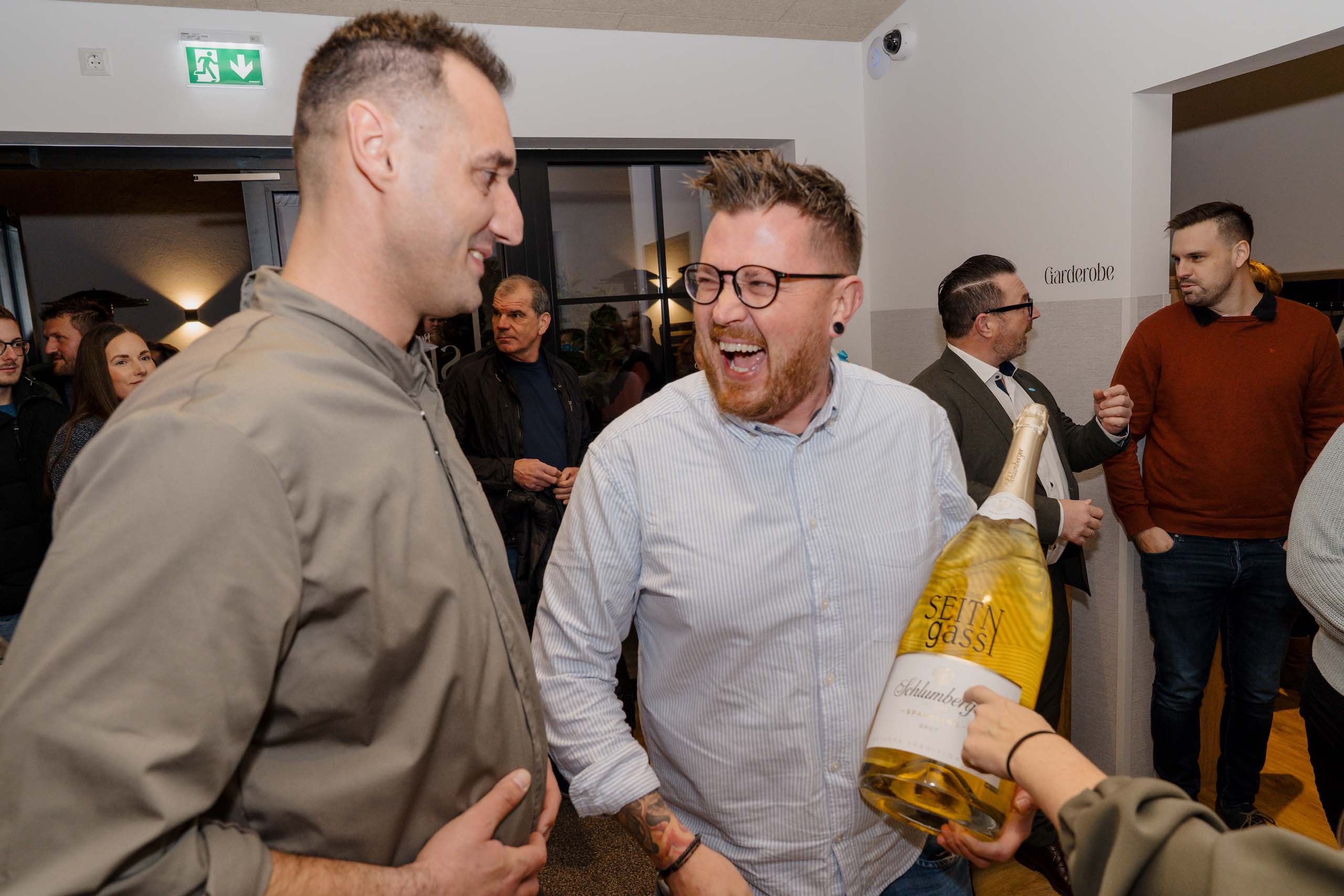 Personen lachen und genießen eine Feier bei einer Hochzeitslocation, halten eine große Flasche Sekt; fröhliche Atmosphäre.