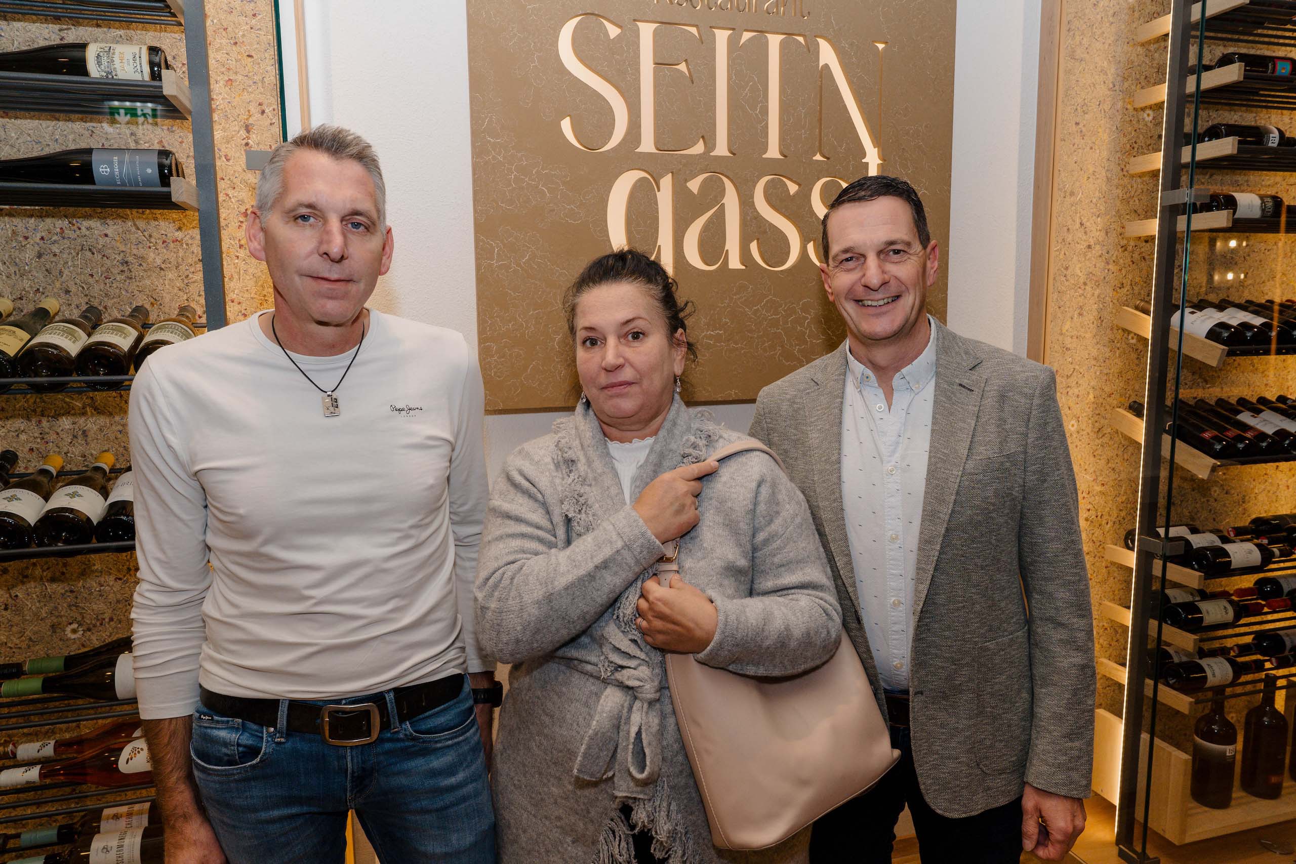 Drei Personen posieren vor einer Weinauswahl im Restaurant, potenzielle Hochzeitstraumlocation für unvergessliche Feiern.