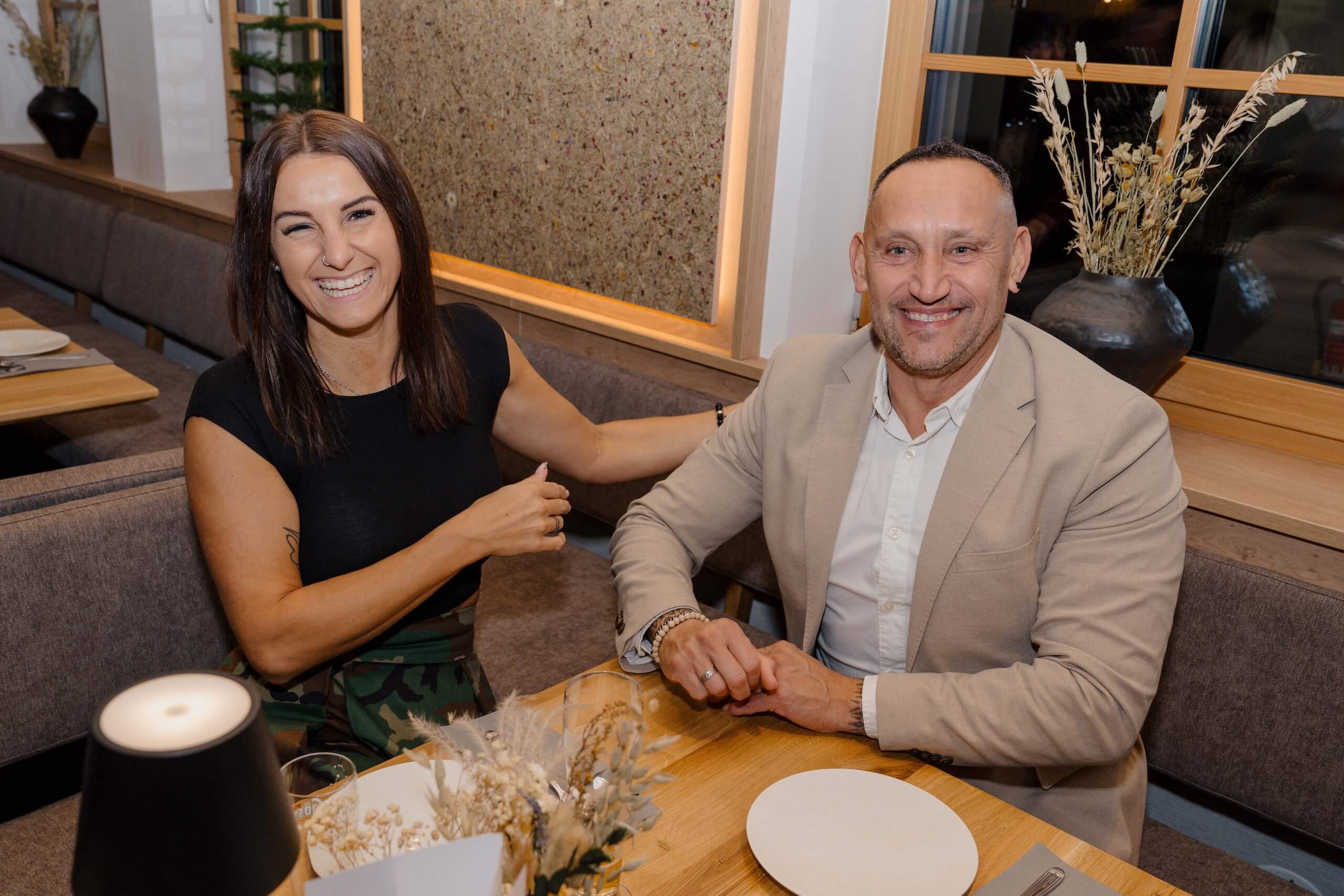 Fröhliches Paar in eleganter Hochzeitslocation, lächelnd am Tisch sitzend. Perfekte Kulisse für den Hochzeitstraum.