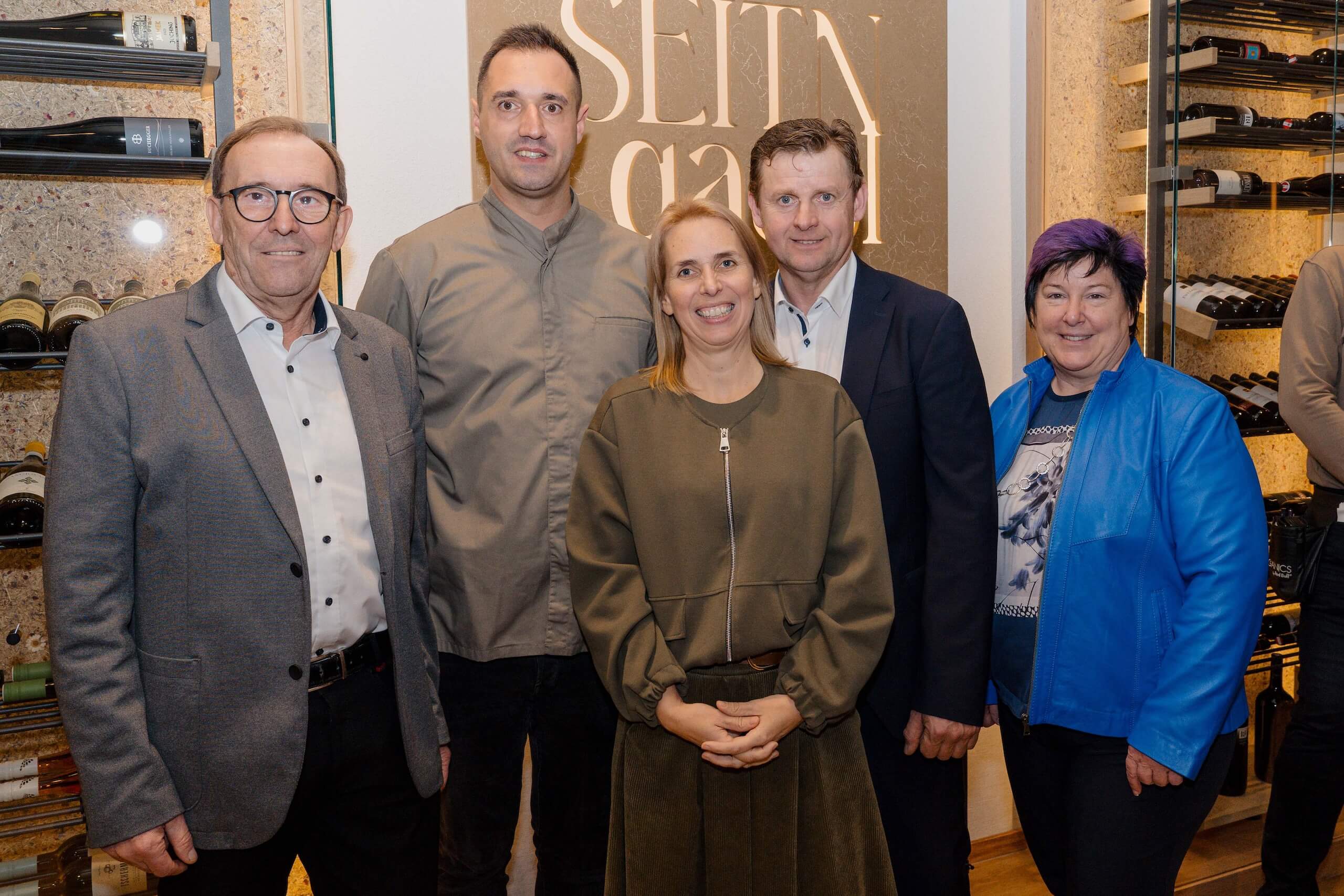 Eine Gruppe von fünf lächelnden Menschen posiert in eleganter Hochzeitslocation vor einem Weinregal.