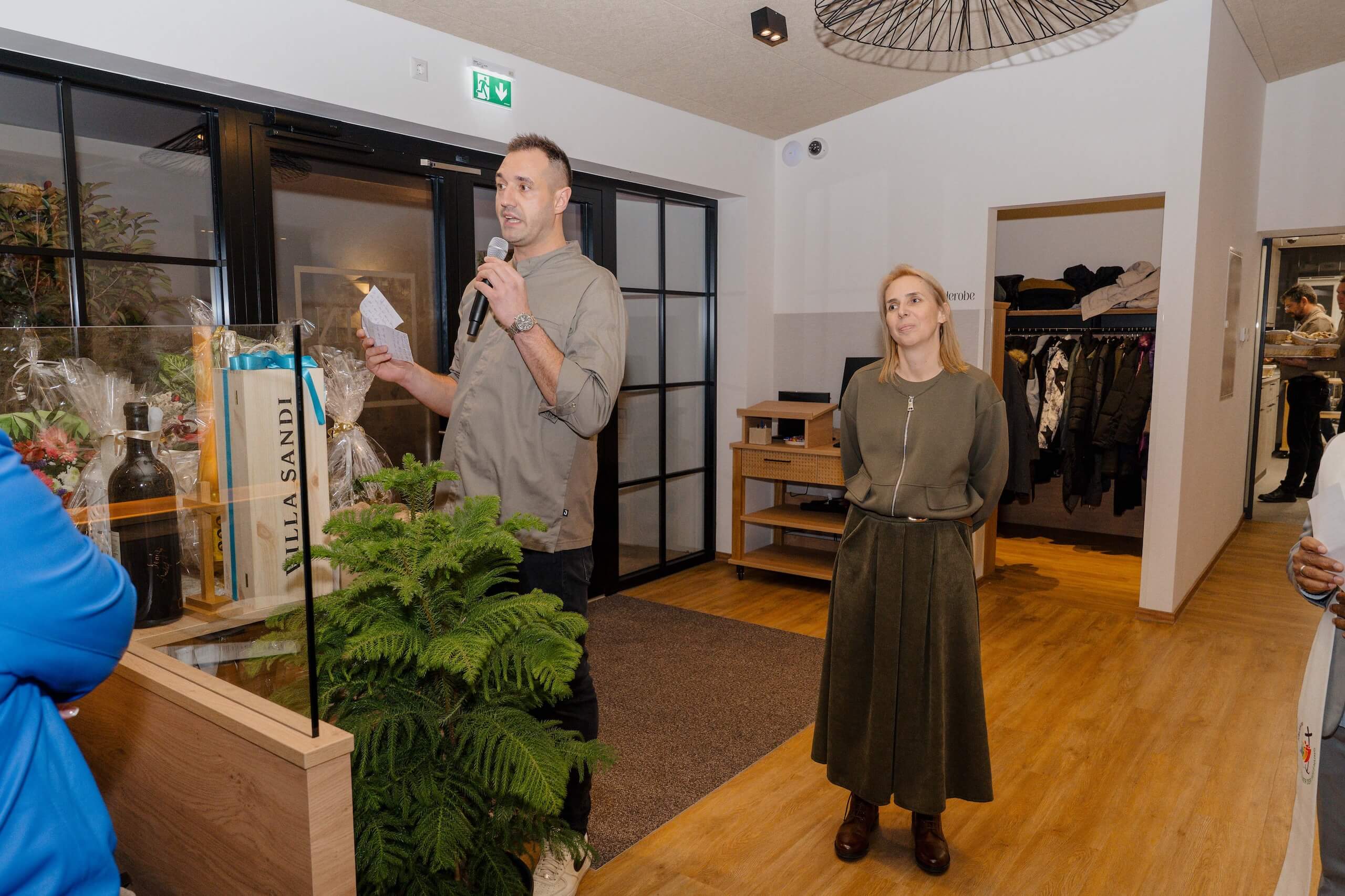 Mann hält Rede bei Hochzeitsempfang in stilvoller Location, dekoriert mit Pflanzen und Geschenkkörben.