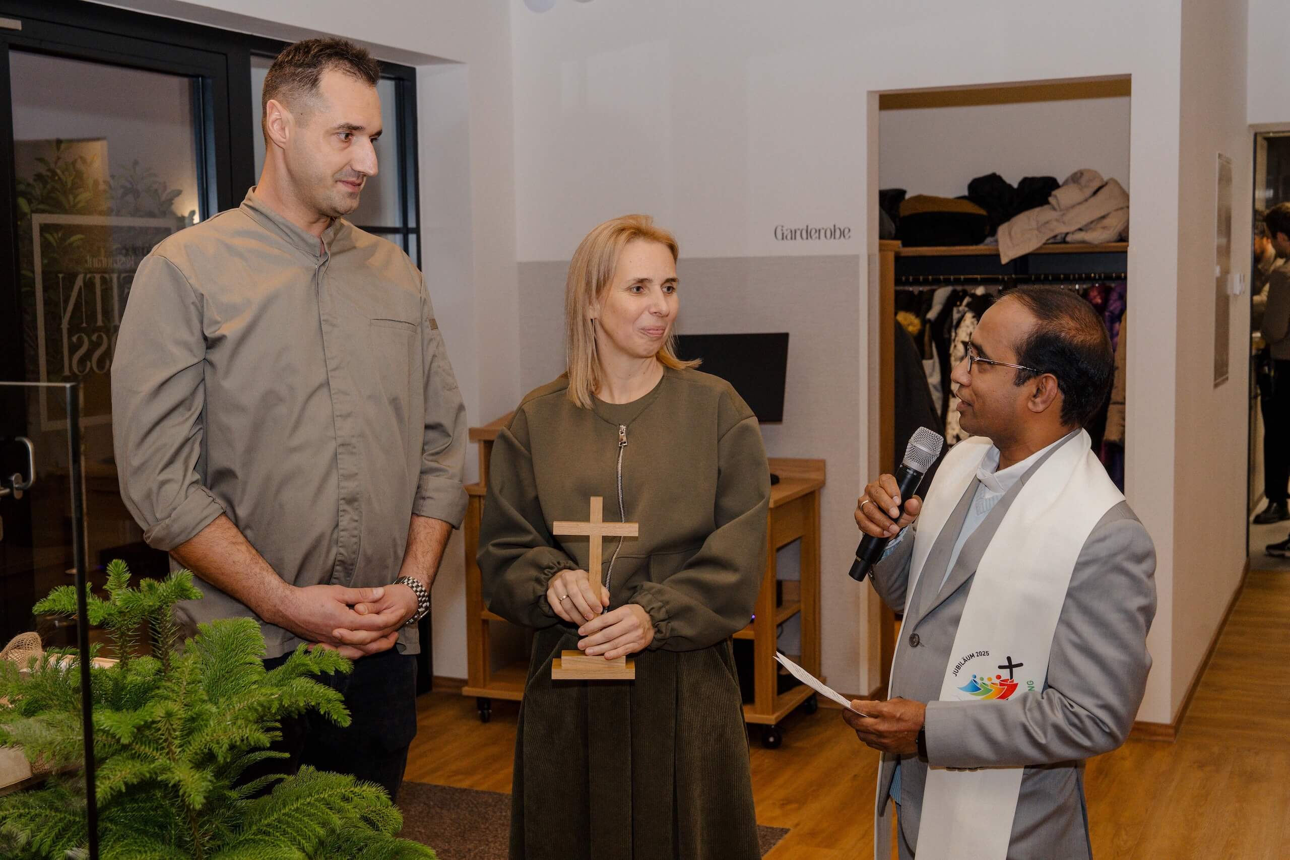 Drei Personen stehen bei einer Hochzeitszeremonie in einer stilvollen Location, jemand spricht ins Mikrofon.