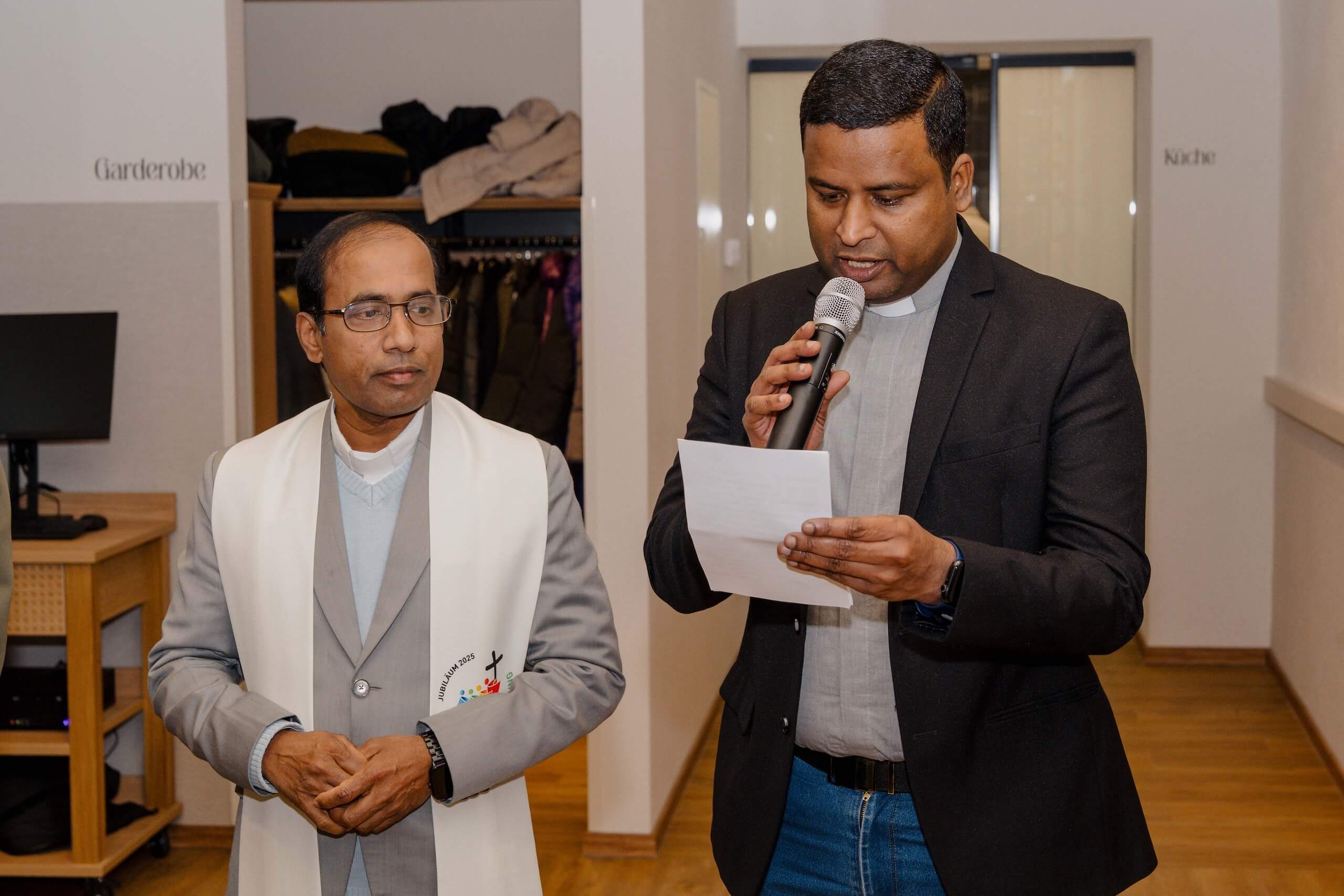 Zwei Männer in Anzügen, einer mit Mikrofon, bereiten sich in einer Hochzeitslocation auf eine Ansprache vor.