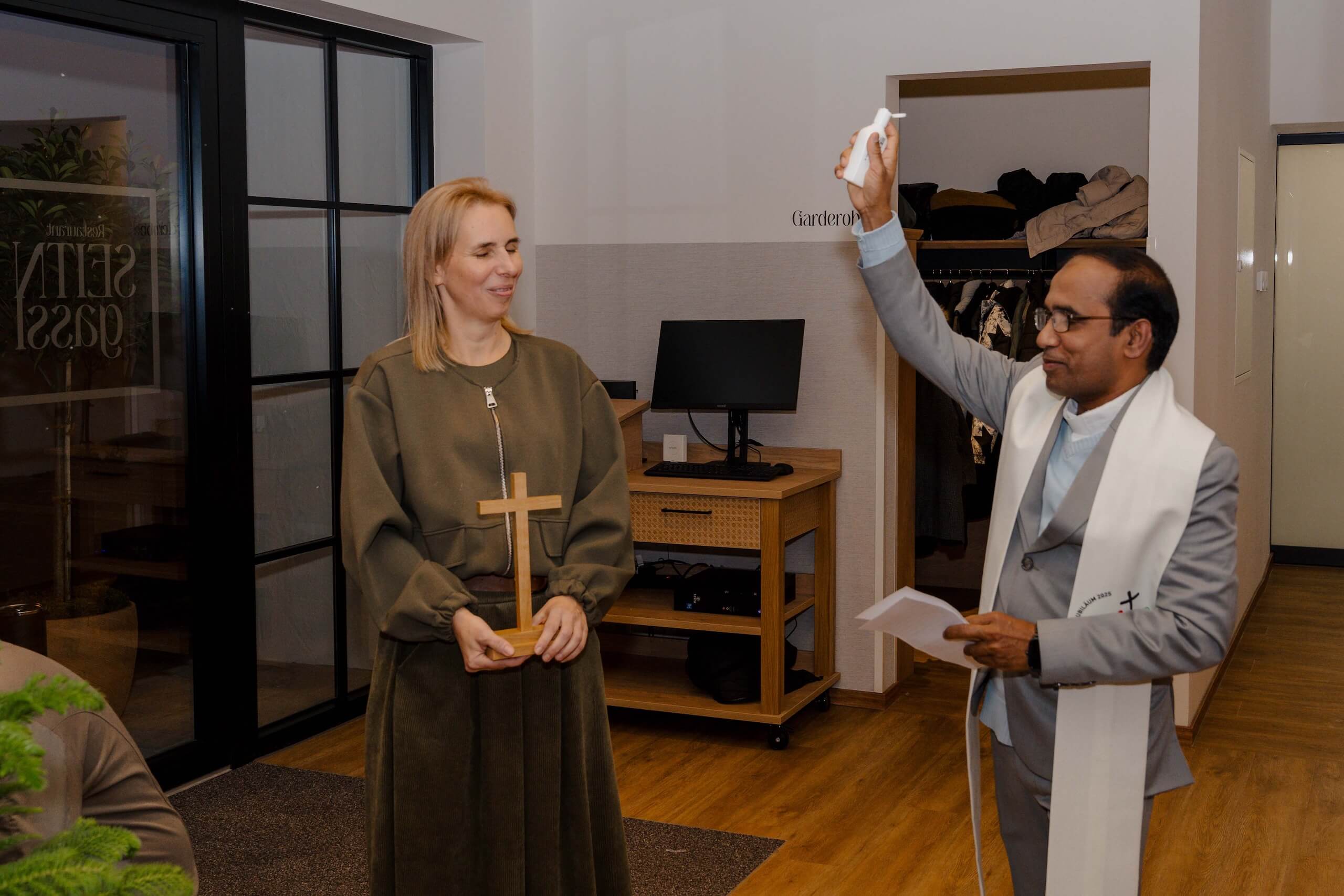 Priester segnet Braut mit Wasser; Braut hält Holzkreuz, lächelt. Innenraum einer eleganten Hochzeitslocation.