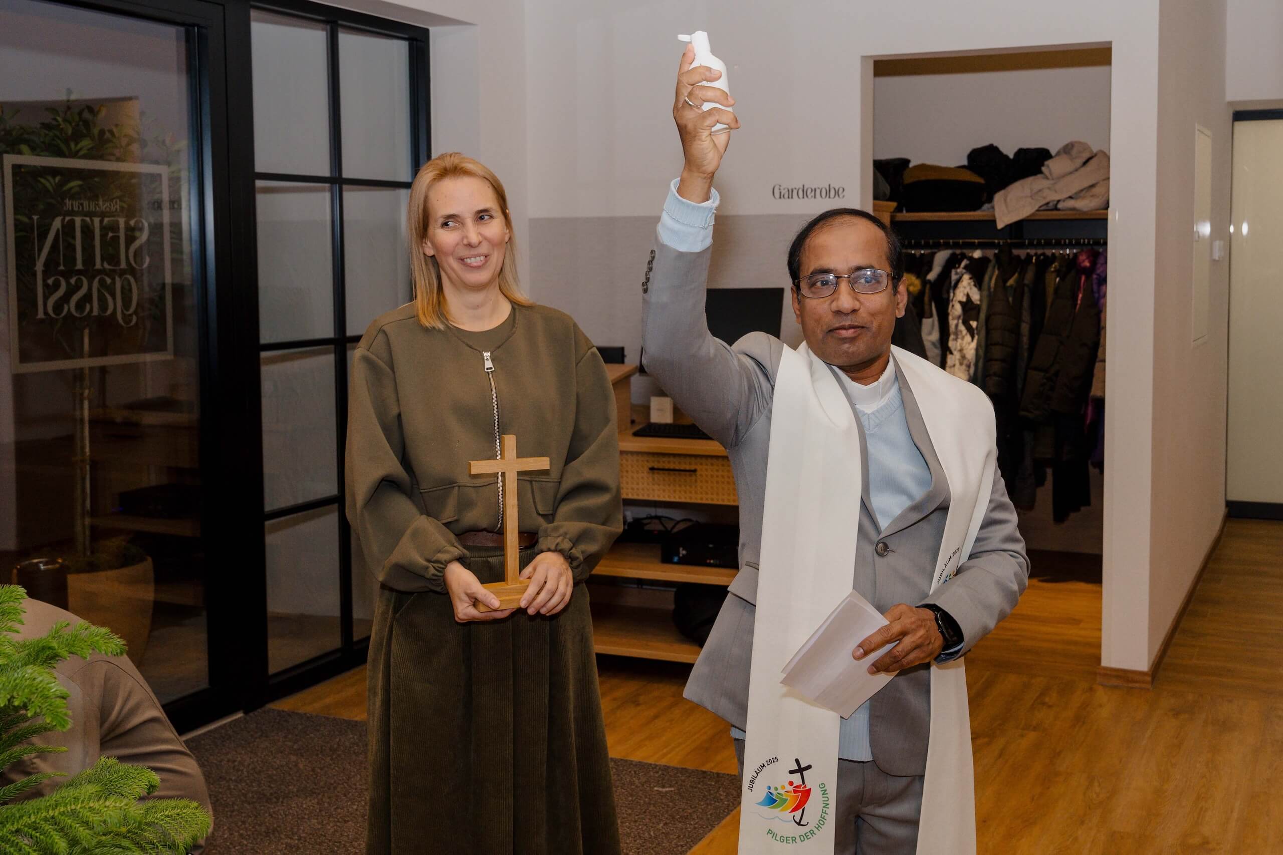 Priester segnet Hochzeitslocation mit Weihwasser, während eine Frau ein Kreuz hält. Perfekter Ort für Traumhochzeiten.