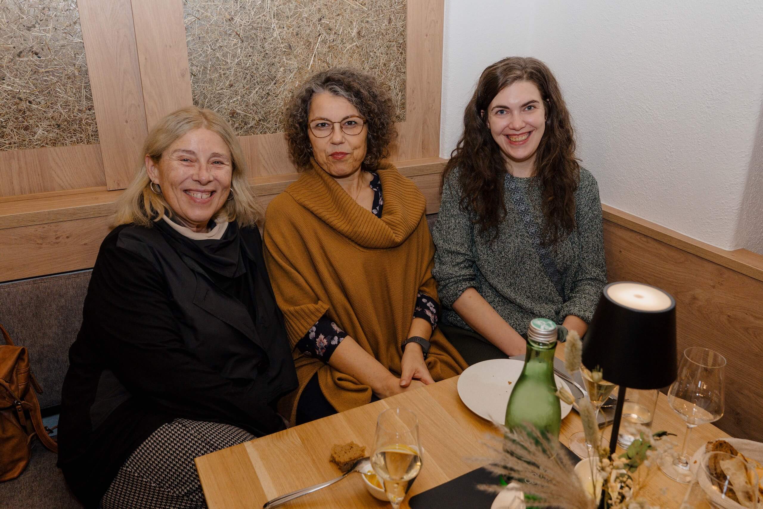 Drei lachende Frauen sitzen fröhlich an einem dekorativen Holztisch, ideal für eine gemütliche Hochzeitslocation.