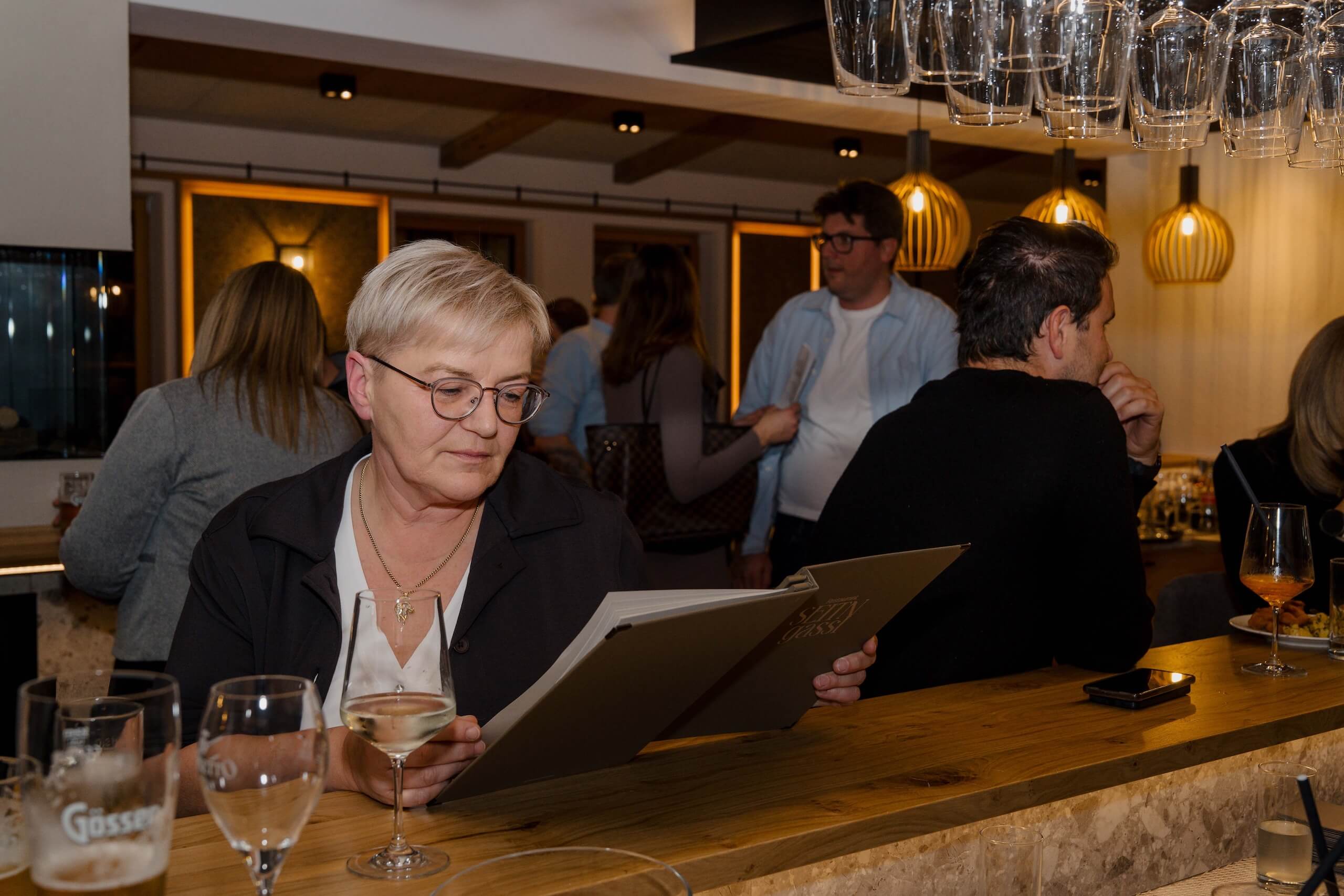 Frau liest Weinkarte an stilvoller Bar in eleganter Hochzeitslocation, Gäste im Hintergrund, warm beleuchtet.