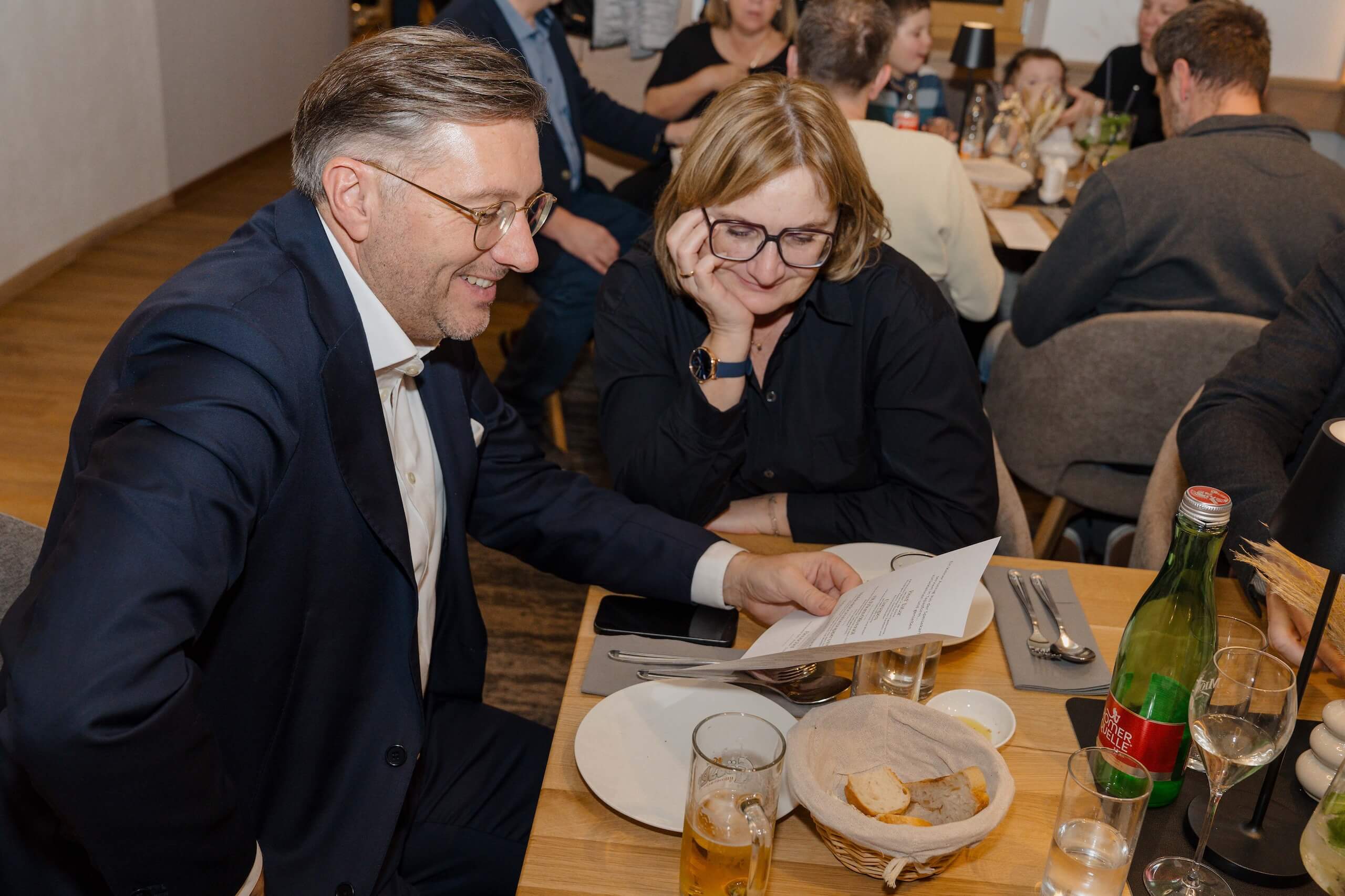 Zwei Personen an einem Tisch besprechen Details bei einer Hochzeit, umgeben von Gästen in einer gemütlichen Hochzeitslocation.