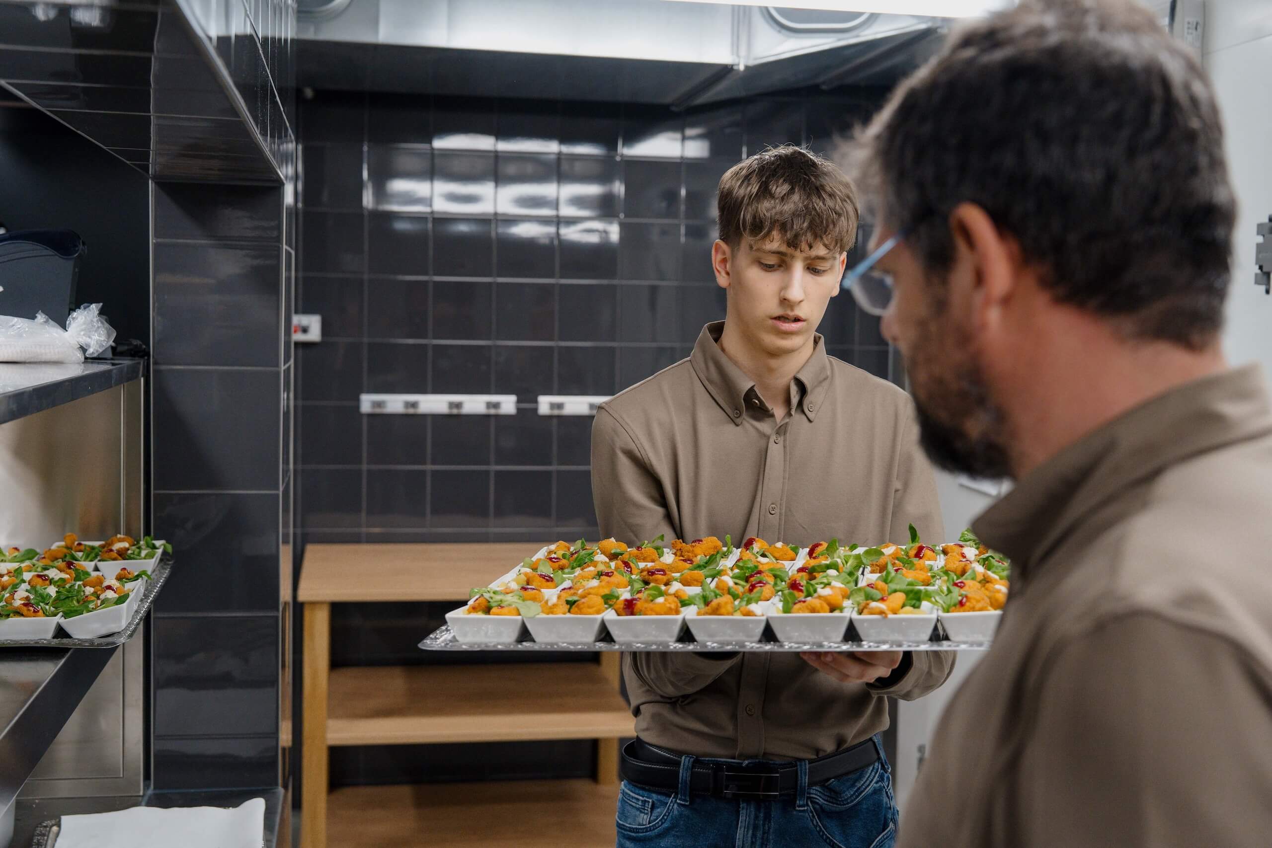 Junger Kellner trägt Tablett mit Vorspeisen für Hochzeitsempfang; Vorbereitung in moderner Hochzeitslocation-Küche.