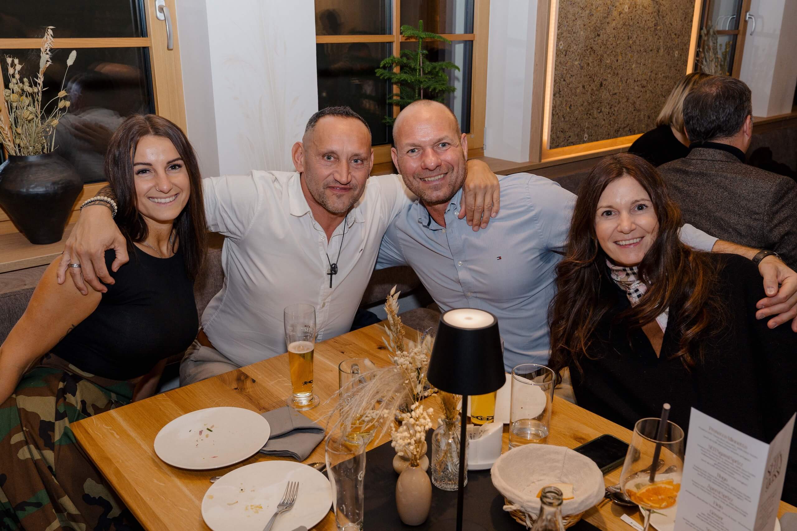 Vier Gäste genießen lächelnd ein Hochzeitsessen in gemütlicher Hochzeitslocation, dekoriert mit eleganten, natürlichen Details.